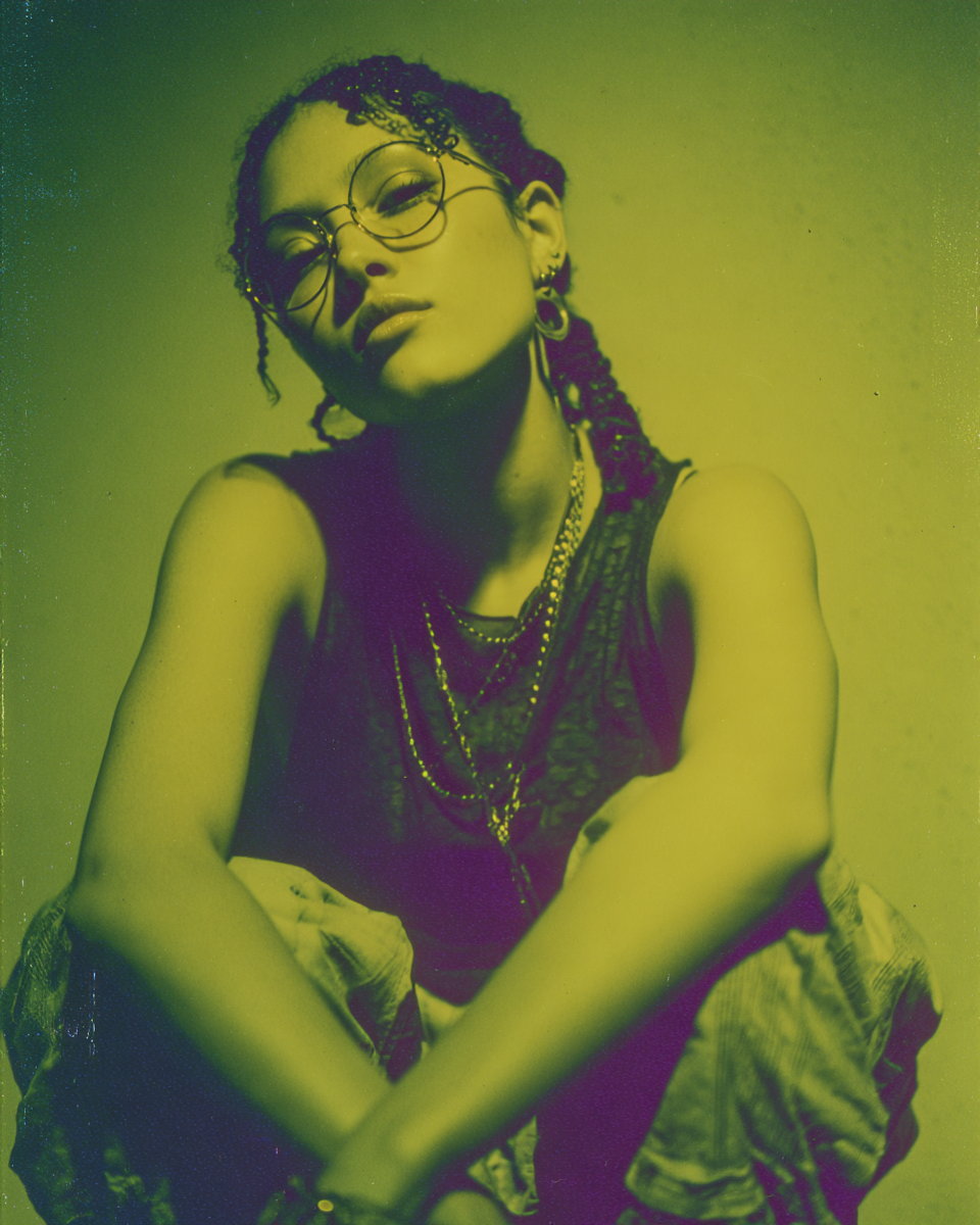 Aiesha cross-processed into acid yellow and magenta, seated with arms folded, layered chains over a ribbed tank, canvas overshirt off the shoulders